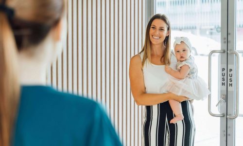 Photo of a smiling blonde haired woman carrying an infant as she arrives for a Brisbane cosmetic dentistry treatment | featured image for Home.